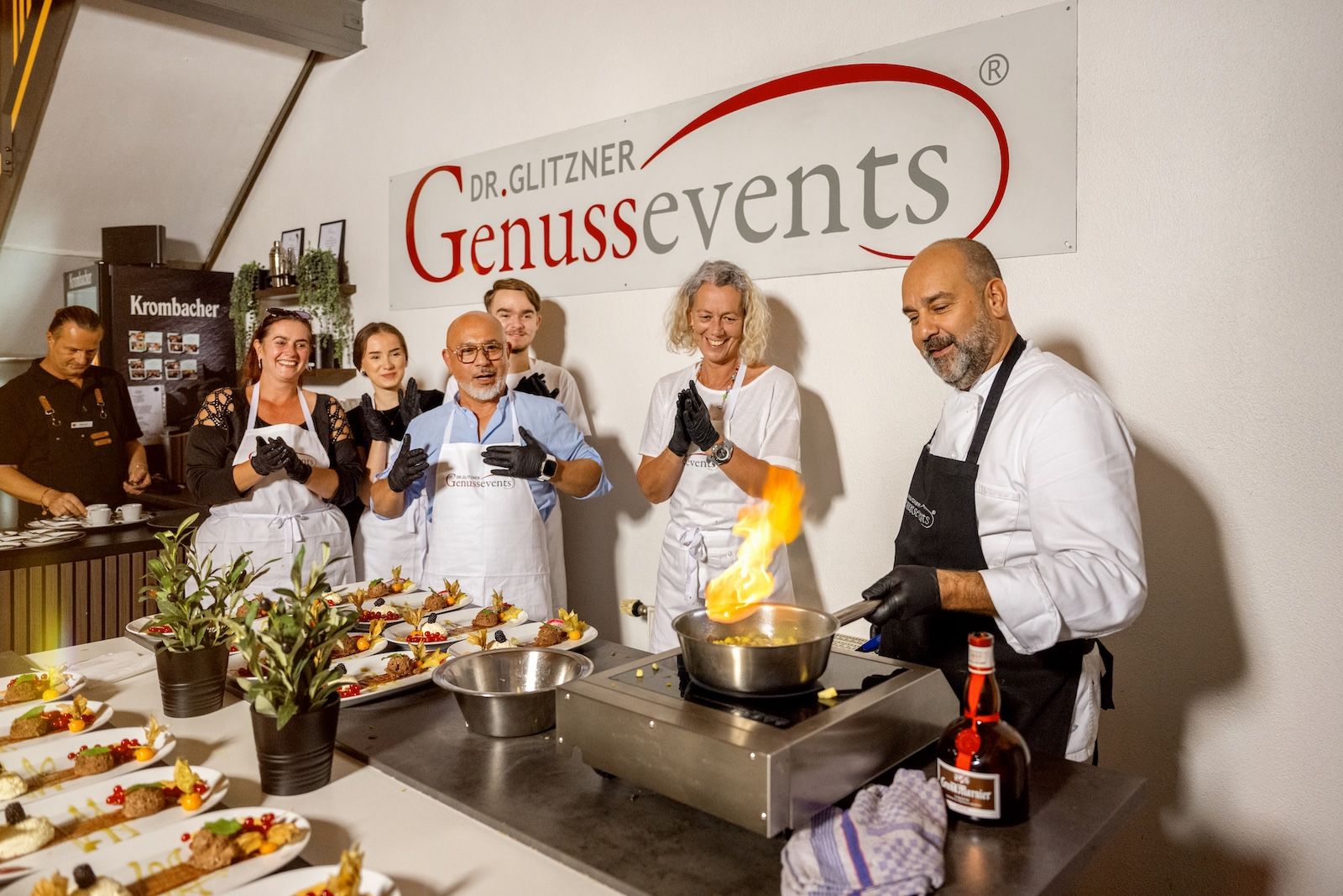 Eine Gruppe von Teilnehmerinnen und Teilnehmern steht in der Kochschule Frankfurt um den Chefkoch, der eine flambierte Speise zubereitet. Die Gäste tragen Schürzen von „GenussEvents“ und applaudieren begeistert