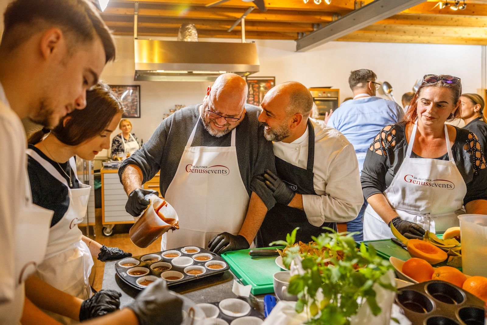 Teilnehmerinnen und Teilnehmer eines Kochkurses in der Kochschule Frankfurt bereiten gemeinsam Desserts zu. Ein Koch unterstützt die Gruppe mit Tipps, während ein Teilnehmer Karamellmasse in Förmchen gießt und konzentriert arbeitet.
