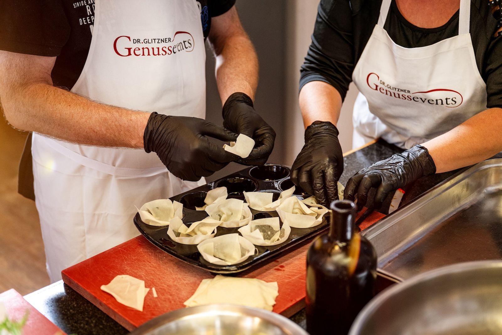 Zwei Personen mit GenussEvents-Schürzen formen Teig in einer Muffinform und bereiten gemeinsam kleine Gebäck- oder Vorspeisenschälchen in der Kochschule Frankfurt zu.